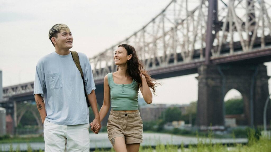 A young couple walking together, smiling and looking at each other, with a bridge and water body in the background.